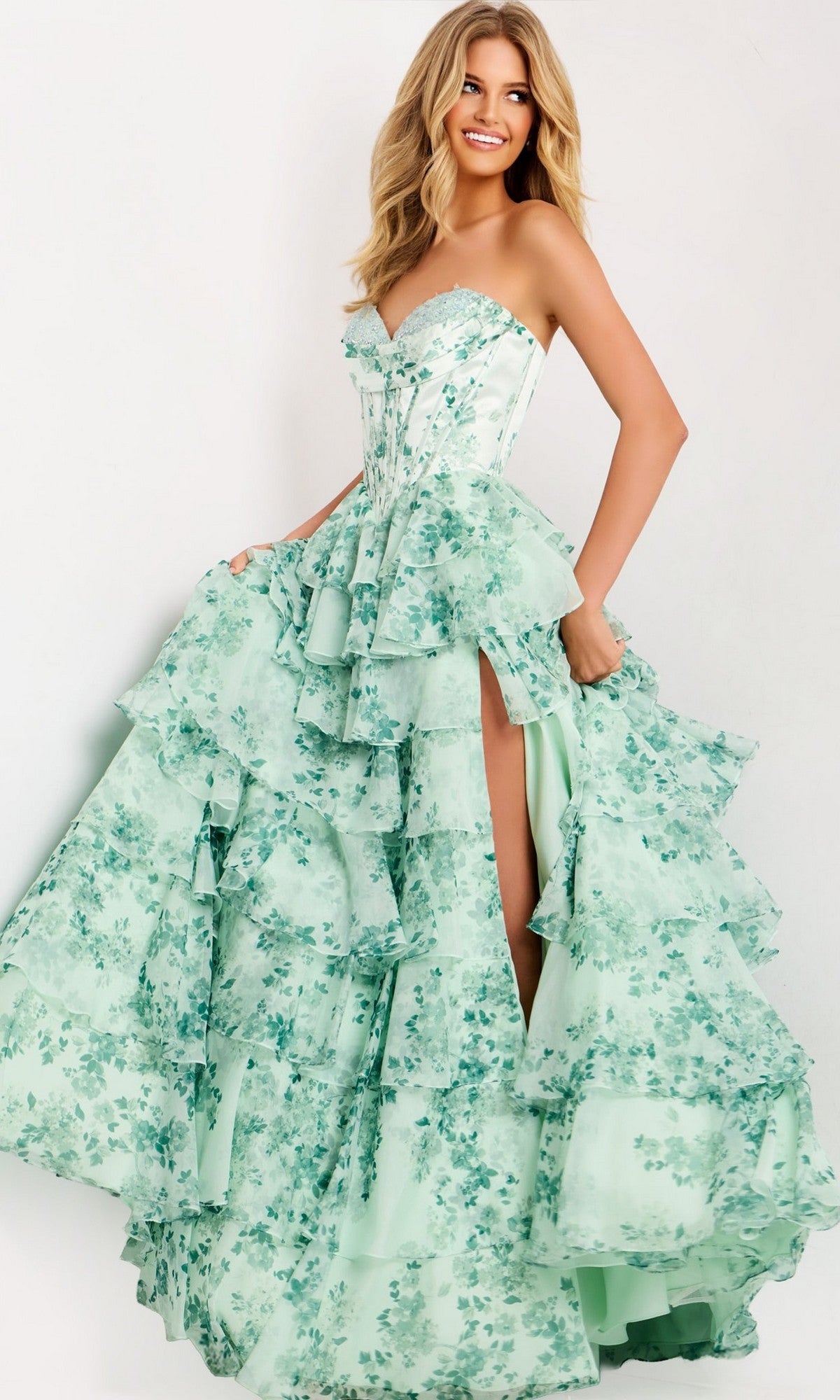A woman wears the Jovani 48772 long prom dress in green/print, featuring a strapless corset bodice, floral pattern, ruffled details, and a high slit, standing against a plain light background.